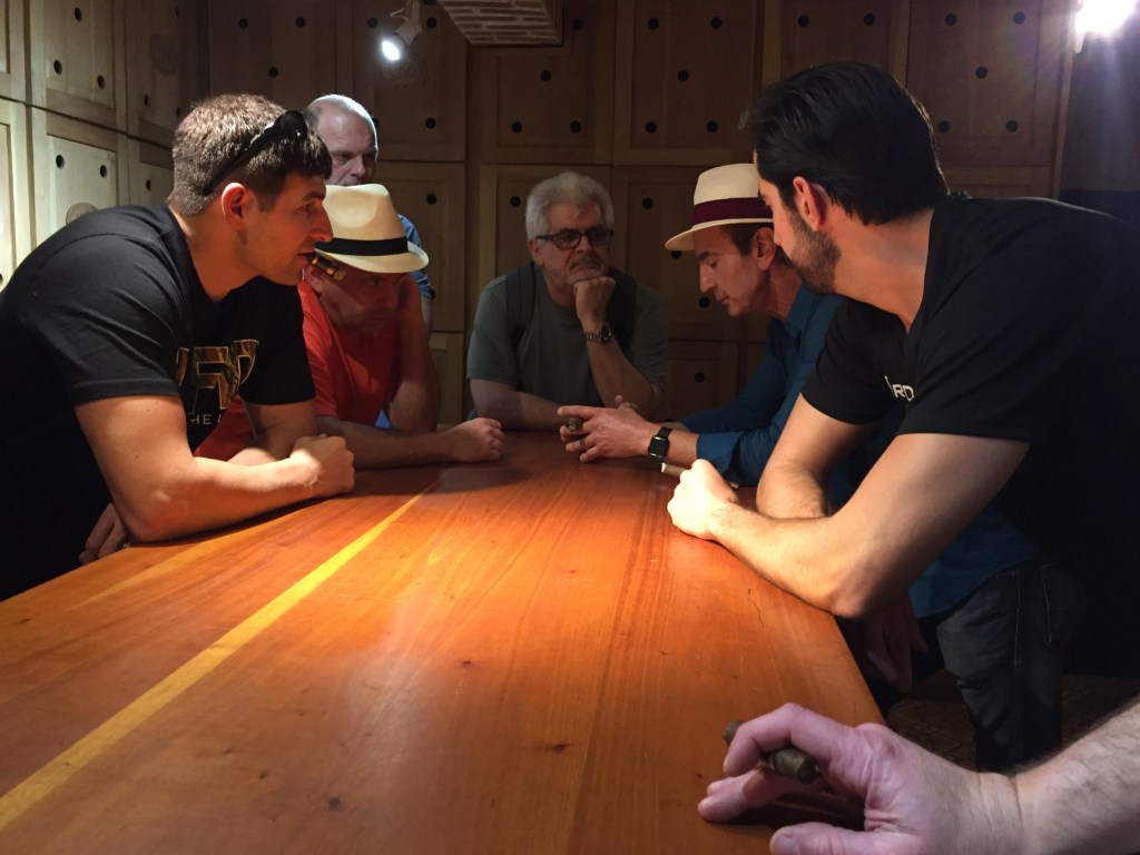 A group of individuals engaged in conversation around a wooden table, some holding cigars, in a cozy, well-appointed cigar lounge setting.