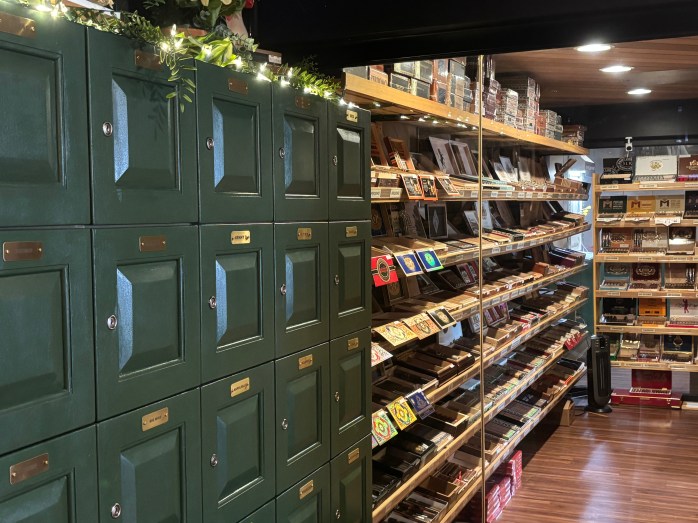An interior view of a retail store featuring green wooden lockers on the left side and shelves filled with various cigar boxes on the right.